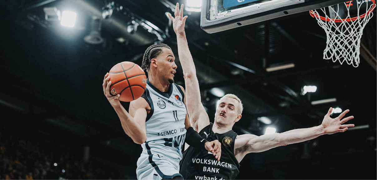 Ein fesselndes Bild zeigt einen spannenden Basketball-Dunk, bei dem ein Spieler in wei em Trikot mit einem kraftvollen Sprung den Ball in der Luft h lt. Der intensive Moment ist eingefroren, mit einem Gegner in schwarzem Trikot, der den Ball blockt, und einer Menge verschwommener, begeisterter Zuschauer im Hintergrund, die die elektrisierende Atmosph re des Sports einfangen.