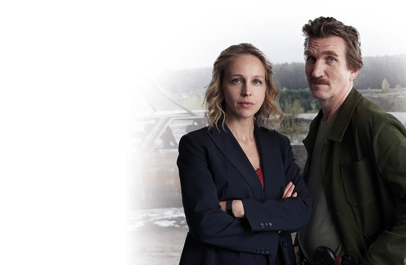 Im Vordergrund stehen eine Frau und ein Mann nebeneinander. Die Frau tr gt einen dunklen Blazer, hat blonde, offene Haare und blickt ernst in die Kamera. Der Mann tr gt eine gr ne Jacke, hat braune Haare und einen Schnurrbart, auch er schaut ernst. Im Hintergrund wird ein stark verbranntes Auto mit einem Kran aus dem Wasser gezogen. Mehrere Personen in wei en Schutzanz gen sowie ein Polizeifahrzeug sind am Einsatzort zu sehen. Die Umgebung wirkt l ndlich, mit Wald und Wasserfl che.