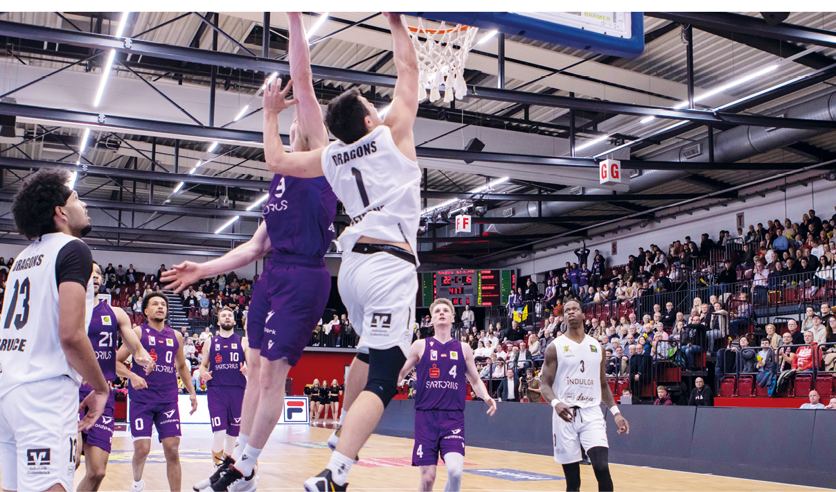 Das Bild zeigt eine Basketballszene in einer Halle mit Zuschauern im Hintergrund. Im Vordergrund sind mehrere Basketballspieler in Aktion zu sehen. Ein Spieler in einem wei en Trikot mit der Nummer 1 springt hoch, um einen Dunking zu machen. Ein anderer Spieler in einem lila Trikot versucht, ihn zu blocken. Weitere Spieler in lila und wei en Trikots stehen auf dem Spielfeld. Die Spieler in lila Trikots tragen Nummern wie 0, 4 und 10. Die Spieler in wei en Trikots tragen Nummern wie 3 und 13.Im Hintergrund sind Zuschauer auf den Trib nen zu sehen. Eine Anzeigetafel zeigt den Spielstand und die verbleibende Zeit an. Die Halle ist mit Lichtern und Werbebannern ausgestattet.