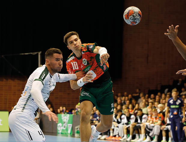 Das Bild zeigt eine dynamische Szene aus einem Handballspiel. Im Mittelpunkt steht ein Spieler in einem orange-gr nen Trikot mit der Nummer 10, der sich in der Luft befindet und im Begriff ist, den Ball zu werfen. Er wird von zwei Verteidigern in wei en Trikots mit gr nen Akzenten flankiert, von denen einer versucht, den Wurf zu blocken. Im Hintergrund ist eine gro e Zuschauermenge zu sehen, die das Spiel aufmerksam verfolgt. Die Halle ist gut beleuchtet und der blaue Bodenbelag sorgt f r einen klaren Kontrast zu den Trikots der Spieler.Das Bild f ngt die Intensit t und Spannung des Handballsports ein und vermittelt einen Eindruck von der Geschwindigkeit und Athletik der Spieler.