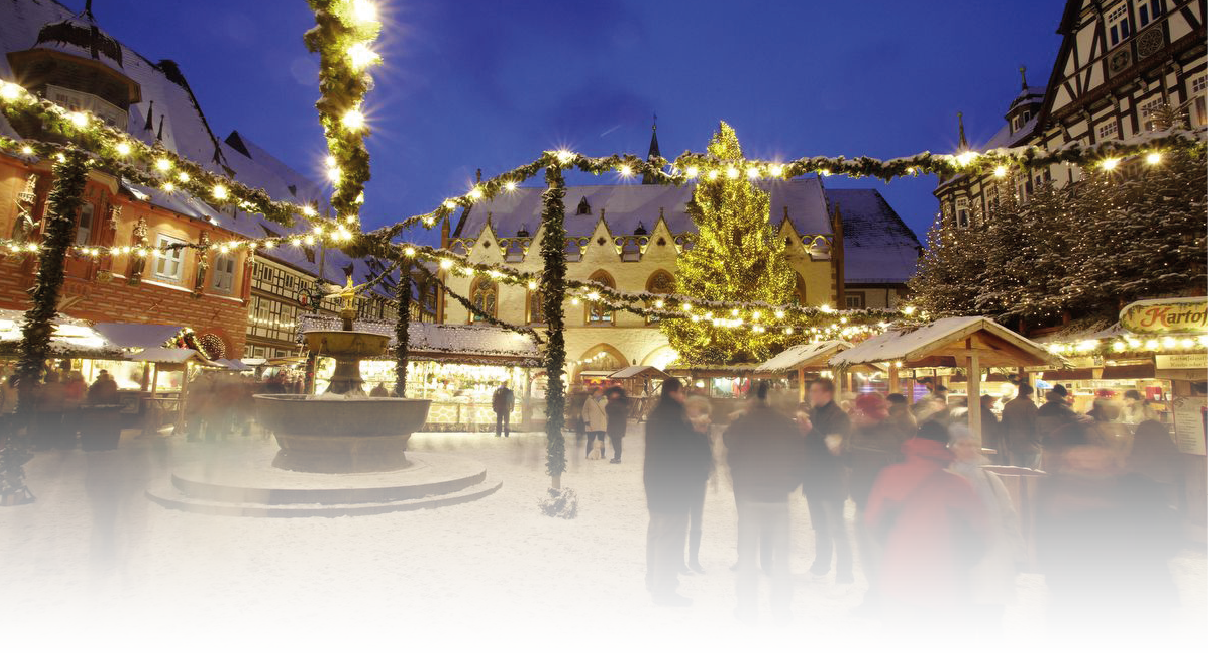 Winterlicher Weihnachtsmarkt auf einem historischen Platz mit verschneitem Boden. Ein gro er Brunnen steht zentral, umgeben von festlich geschm ckten St nden. ber dem Platz h ngen Girlanden mit warmen Lichterketten. Im Hintergrund sind Fachwerkh user und ein gro es Geb ude mit beleuchtetem Weihnachtsbaum zu sehen. Menschen bewegen sich zwischen den St nden.