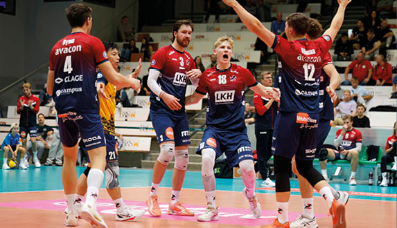 Mehrere Volleyballspieler in rot-blauen Trikots stehen auf einem Hallen-Spielfeld und feiern gemeinsam. Ein Spieler hebt beide Arme in die Luft, w hrend die anderen sich in der Mitte des Feldes versammeln. Im Hintergrund sind Zuschauer auf Trib nen zu sehen. Das Spielfeld hat eine t rkisfarbene Fl che mit einem orangefarbenen Bereich nahe des Netzes.