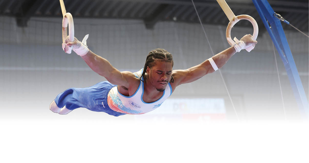 Ein Turner f hrt eine bung an den Ringen aus, mit ausgestreckten Armen in einer horizontalen Position. Er tr gt ein blaues Trikot mit wei en und orangefarbenen Akzenten sowie blaue Turnhose. Die H nde sind mit wei en Bandagen umwickelt, und die Ringe sind an Gurten befestigt. Im Hintergrund ist eine Anzeigetafel mit Punkten und Text zu sehen, sowie ein Netz an der Hallendecke.