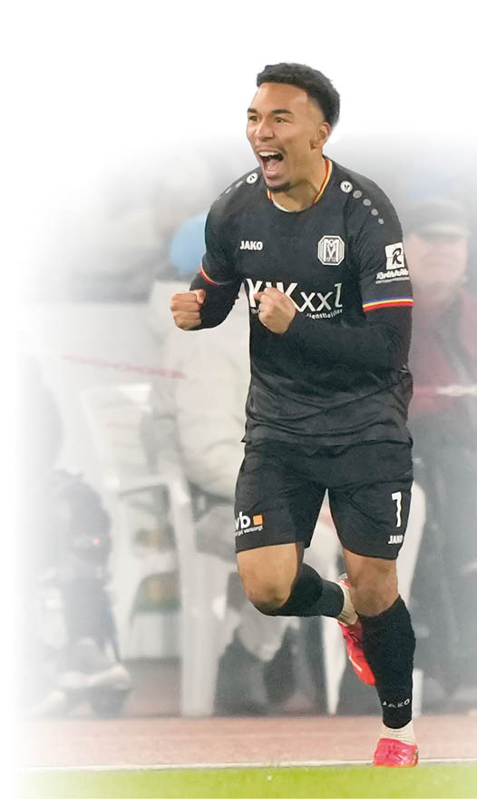 Der Fu ballspieler Oliver Schmitt in schwarzem Trikot mit der Nummer 3 und roten Schuhen l uft auf dem Spielfeld. Das Trikot tr gt mehrere Sponsorenlogos. Im Hintergrund sind Zuschauer auf wei en Plastikst hlen und eine Absperrung mit rot-wei em Band zu sehen.