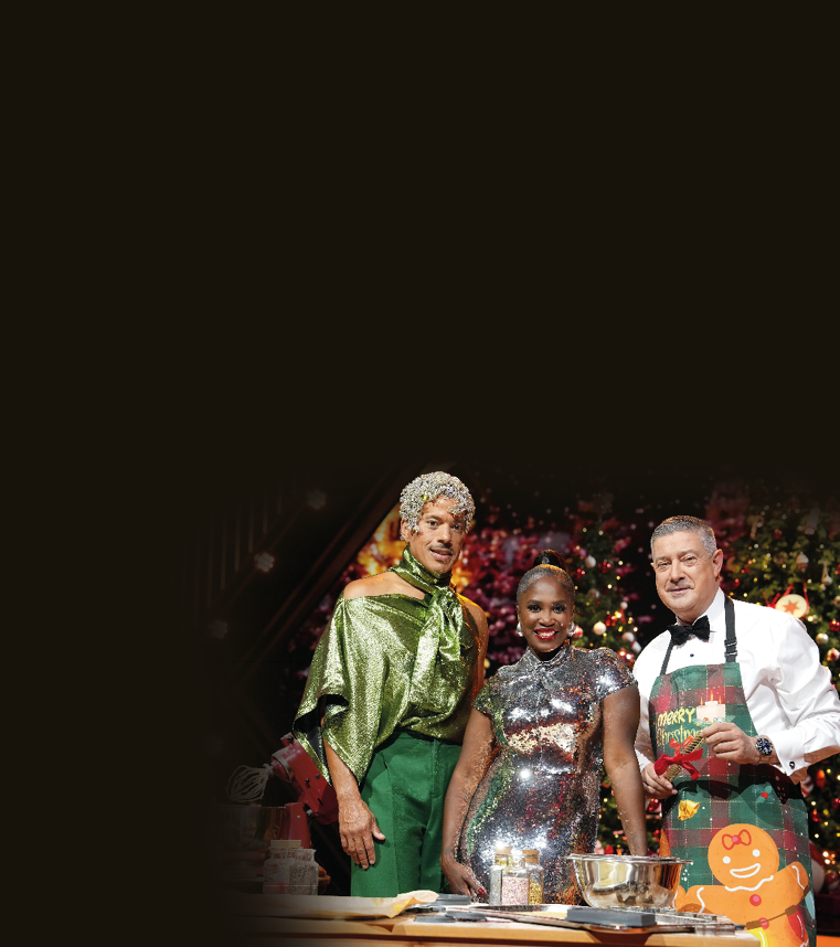 Auf dem Bild stehen drei Personen in festlicher Studio-Kulisse vor weihnachtlich geschm ckten Tannenb umen und berufenen Lichtern. Alle drei sind elegant bzw. auff llig gekleidet: • Der Mann links tr gt ein gl nzendes, gr nes Oberteil mit Schal-Element und eine gr ne Hose; sein Kopf ist mit silbrigem Glitzerschmuck dekoriert.  • Die Frau in der Mitte tr gt ein figurbetontes Kleid aus silbernen, stark reflektierenden Pailletten.  • Der Mann rechts tr gt Hemd, Fliege und eine weihnachtliche Sch rze mit „Merry Christmas“-Aufdruck.  Vor ihnen befindet sich eine Arbeitsfl che mit Back- und Kochutensilien wie Sch sseln, Gew rzgl sern und einem Mixer.   