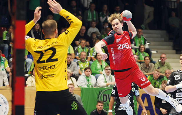 Handballszene in einer Halle: Ein Spieler in Rot springt mit dem Ball in der Hand in Richtung Tor, der Torwart in Gelb steht mit erhobenen Armen bereit. Weitere Spieler in schwarzen Trikots und Zuschauer im Hintergrund.