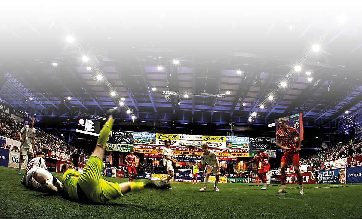 Hallenfu ballspiel aus der Perspektive hinter dem Tor: Ein Torwart in Gelb liegt auf dem Boden, Spieler in Rot und Grau bewegen sich vor dem Tor. Die Halle ist voll besetzt, mit auff lliger blauer Beleuchtung und zahlreichen Werbebannern.