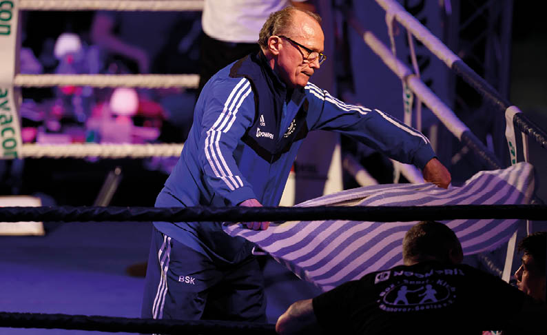 Eine Person in blauer Trainingsjacke steht in einem Boxring und h lt ein gestreiftes Handtuch. Im Vordergrund sind zwei weitere Personen am Ringrand zu sehen, eine davon tr gt ein schwarzes T-Shirt mit wei em Aufdruck. Im Hintergrund sind die Seile des Rings und unscharfe Zuschauer erkennbar, die Szene ist in violettem Licht beleuchtet