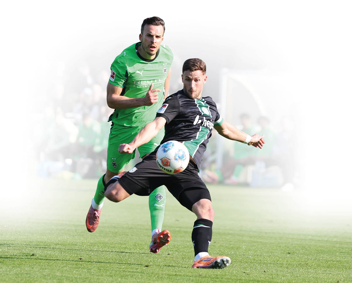 Fu ballszenen auf einem Spielfeld: Ein Spieler in schwarzem Trikot mit gr nem Streifen kontrolliert den Ball, w hrend ein Spieler in gr nem Trikot versucht, den Ball zu erreichen. Im Hintergrund sind weitere Spieler in gr nen Trikots auf einer Bank sowie Zuschauer und ein Trainerbereich mit einer Ersatzbank zu sehen. Die Szene findet bei sonnigem Wetter auf einem gepflegten Rasenplatz statt.