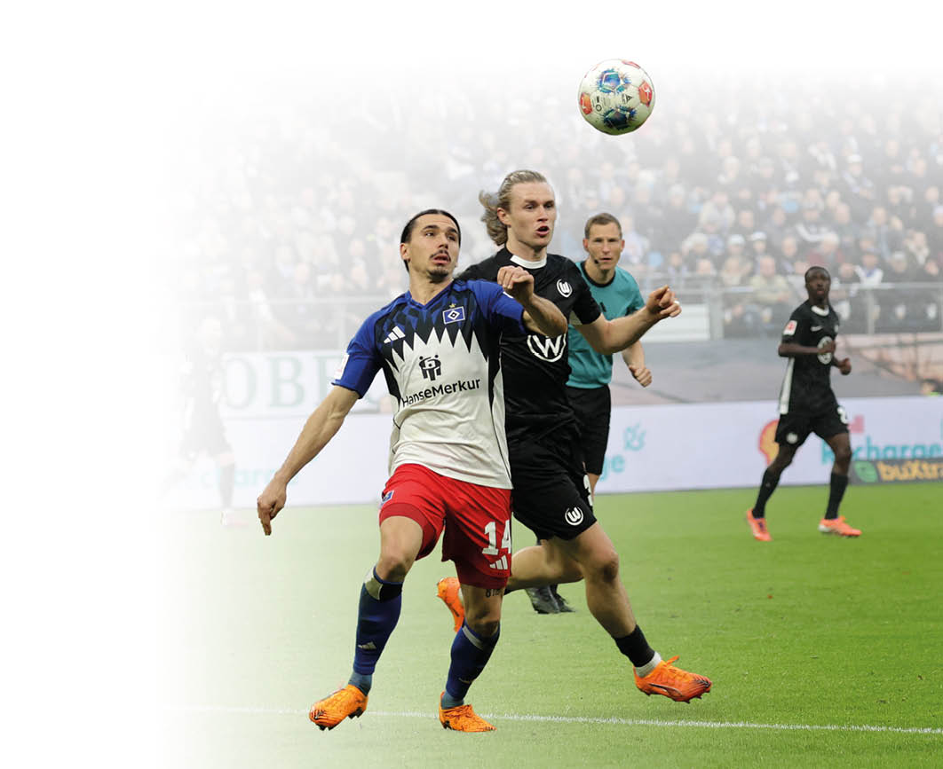 Zwei Fu ballspieler k mpfen im Stadion um einen hoch springenden Ball; im Vordergrund stehen sich ein Spieler im blau-wei -roten Trikot und ein Spieler im schwarzen Trikot gegen ber, w hrend im Hintergrund weitere Spieler und eine dicht besetzte Trib ne zu sehen sind.