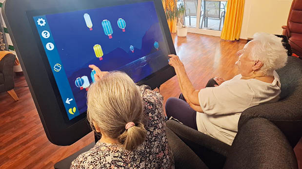 Zwei ltere Personen sitzen in einem hellen Gemeinschaftsraum und interagieren gemeinsam mit einem gro en digitalen Aktiv-Tisch, auf dessen Bildschirm ein farbenfrohes Spiel mit schwebenden Laternen zu sehen ist.