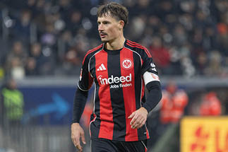 Mann in schwarzem Mantel mit erhobenen H nden vor unscharfem Hintergrund mit Menschen und rotem Banner