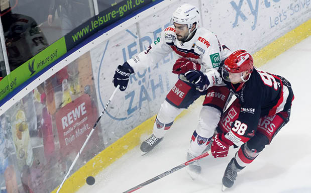 Zwei Eishockeyspieler liefern sich an der Bande ein intensives Duell um den Puck, w hrend sie mit ihren Schl gern um die Kontrolle k mpfen und Eis aufwirbelt.