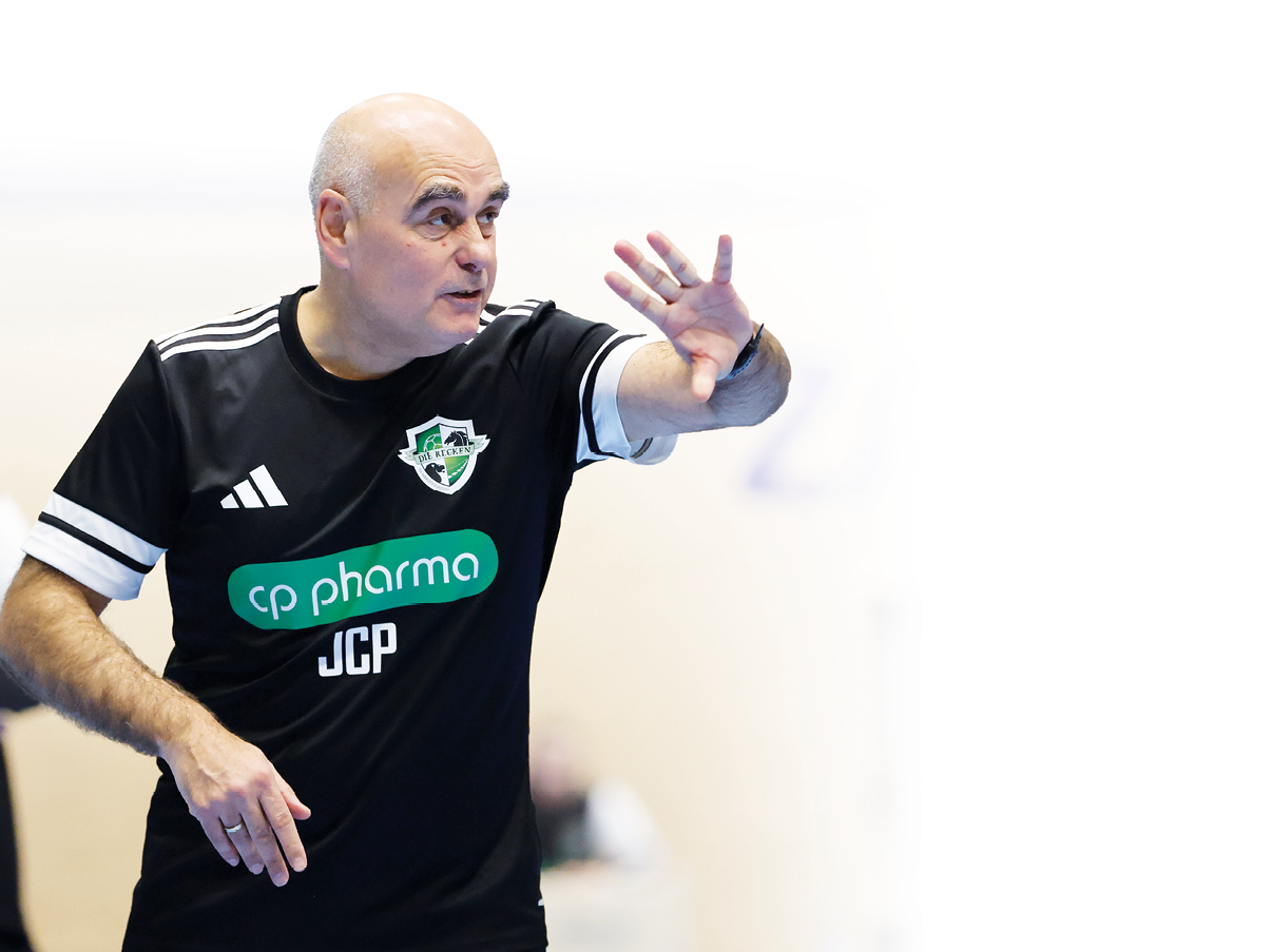 Ein Handballtrainer in schwarzer Sportkleidung steht in einer Sporthalle am Spielfeldrand, hebt eine Hand mit ausgestreckten Fingern und gibt gestenreich Anweisungen. Auf seinem Trikot sind ein Vereinslogo sowie ein Sponsorenschriftzug zu sehen; im Hintergrund stehen weitere Teammitglieder und Hallenbeschilderungen.