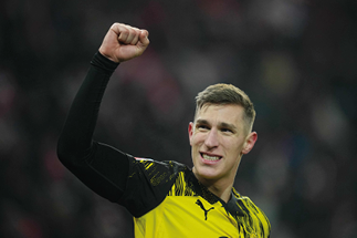 Mann in schwarzem Mantel mit erhobenen H nden vor unscharfem Hintergrund mit Menschen und rotem Banner