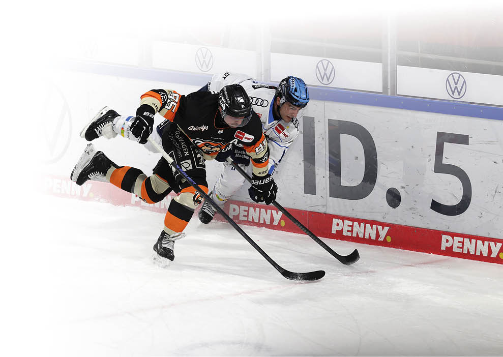 Zwei Eishockeyspieler k mpfen dynamisch an der Bande um den Puck. Einer der Spieler tr gt ein schwarzes Trikot, der andere ein wei es. Beide beugen sich stark nach vorn, w hrend ihre Schl ger den Puck verfolgen. Im Hintergrund sind Werbebanden und das Eisstadion zu sehen.