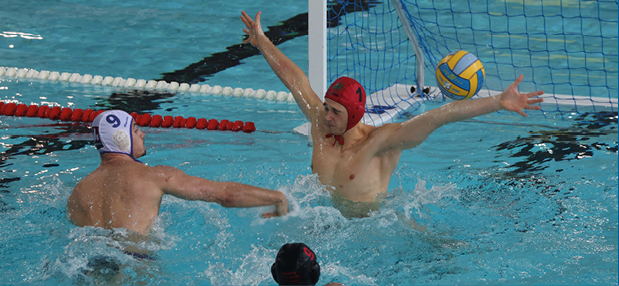 Ein Wasserballtorwart mit roter Badekappe springt im Schwimmbecken mit weit ausgebreiteten Armen vor dem Tor hoch, um einen Wurf abzuwehren. Ein Gegenspieler mit wei er Kappe steht nah vor ihm und versucht zu schie en, w hrend ein weiterer Spieler im Vordergrund ins Geschehen schwimmt. Der gelb-blaue Ball fliegt dicht am Tor vorbei, Wasser spritzt um die Spieler herum.