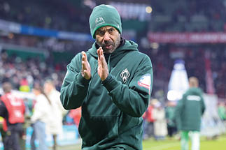 Mann in schwarzem Mantel mit erhobenen H nden vor unscharfem Hintergrund mit Menschen und rotem Banner