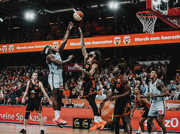 Ein actionreicher Moment eines Basketballspiels: Eine Person im hellen Trikot springt in der N he des Korbs hoch und wirft den Ball einh ndig in Richtung Ring. Mehrere Personen im dunklen Trikot versuchen, den Wurf zu blocken. Weitere Spielende stehen in unmittelbarer N he, w hrend im Hintergrund eine gro e, gef llte Zuschauertrib ne und leuchtende Werbebanden zu sehen sind.