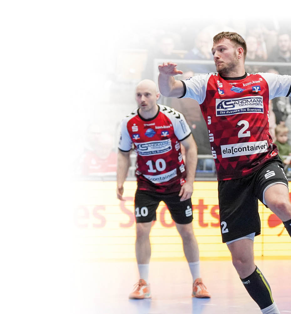 Eine Person im rot schwarzen Handballtrikot springt mit ausgestrecktem Arm und h lt den Ball bereit zum Wurf. Mehrere Spielende in gelben oder rot schwarzen Trikots befinden sich in unmittelbarer N he und beobachten die Aktion oder bereiten sich auf die Abwehr vor. Im Hintergrund sind Trib nen mit Publikum und eine leuchtende Werbebande am Spielfeldrand zu sehen.