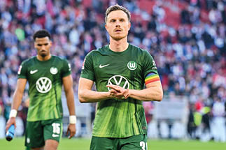 Zwei Fu ballspieler in gr nen Trikots mit Volkswagen-Logo, einer tr gt ein Regenbogenarmband am linken Arm, im Hintergrund Zuschauer im Stadion
