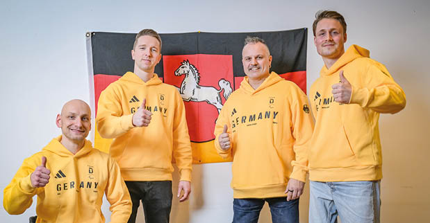 Vier Personen stehen und sitzen vor einer Wand, an der eine Flagge mit einem wei en Pferd auf rotem Hintergrund befestigt ist. Alle tragen gelbe Hoodies mit der Aufschrift „Germany“ und zeigen einen Daumen nach oben. Eine Person sitzt in einem Rollstuhl, w hrend die anderen daneben stehen.