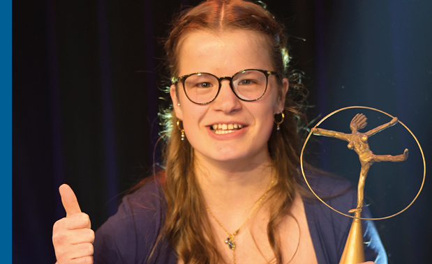 Eine Frau in festlicher Abendkleidung h lt eine goldfarbene Troph e mit einer filigranen Figuren-Skulptur darauf in der einen Hand und zeigt mit der anderen Hand einen erhobenen Daumen. Der Hintergrund ist dunkel und von B hnenlicht beleuchtet.
