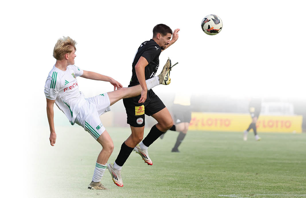 Eine Szene aus einem Fu ballspiel auf einem Rasenplatz. Eine Person in einem wei en Trikot versucht, mit einem ausgestreckten Bein den Ball zu spielen, w hrend eine Person in einem schwarzen Trikot gleichzeitig zum Ball springt. Der Ball befindet sich in der Luft ber den beiden Spielerpersonen. Im Hintergrund stehen weitere Spielerpersonen sowie eine Schiedsrichterperson.