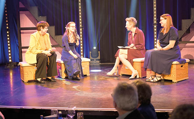 Vier Personen sitzen auf kleinen Holzb nken auf einer B hne im Gespr chskreis. Die B hne ist mit Scheinwerfern ausgeleuchtet, im Vordergrund sind Zuschauer und Tische sichtbar. Die Atmosph re wirkt wie eine moderierte Gespr chsrunde w hrend einer Gala oder Preisverleihung.
