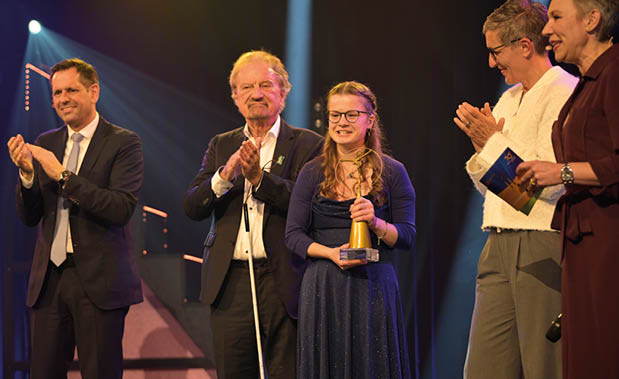 Auf einer festlich beleuchteten B hne stehen f nf Personen nebeneinander. Die Person in der Mitte h lt einen goldfarbenen Award in den H nden und tr gt ein dunkelblaues, bodenlanges Kleid. Die Personen links und rechts davon applaudieren. Eine Person auf der rechten Seite h lt ein Moderationskarten-Set. Spotlights und dunkler B hnenhintergrund schaffen eine feierliche Atmosph re.
