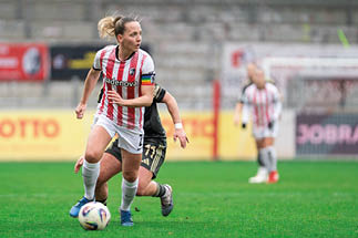Eine Spielerin in wei er Hose und rot‑wei gestreiftem Trikot dribbelt den Ball vorn auf dem gr nen Rasen, w hrend eine Gegenspielerin in dunklem Trikot knapp hinter ihr versucht, sie zu stoppen. Die vordere Spielerin tr gt ein buntes Kapit nsband am linken Oberarm und blaue Schuhe; das Ballger t liegt knapp vor ihren F  en. Im Hintergrund sieht man unscharf eine Torlinie und eine weitere Mitspielerin sowie leere Stadionr nge. Die Szene wirkt dynamisch und zeigt eine Spielsituation im Frauenfu ball. Gesichter sind unscharf oder unkenntlich; keine weiteren Personen oder Orte sind eindeutig identifizierbar.