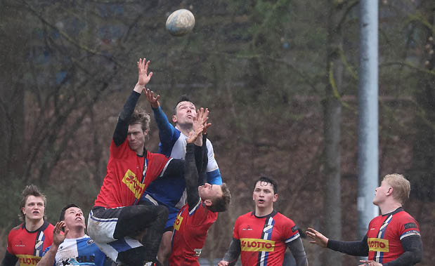 Mehrere Rugbyspieler in roten und blau-wei en Trikots springen gleichzeitig in die Luft, um einen hohen Ball zu fangen. Die Szene wirkt dynamisch und k rperbetont, w hrend weitere Spieler gespannt zuschauen. Der Boden ist matschig, und die Aktion findet auf einem Au enspielfeld statt.