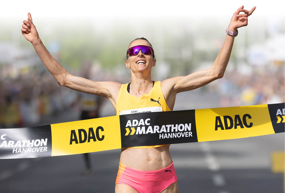 L uferin jubelt mit erhobenen Armen, w hrend sie beim ADAC Marathon Hannover das Zielband durchl uft. Sie tr gt ein gelbes Laufoberteil, pinke Shorts und Sportsonnenbrille.