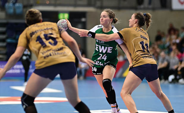 Handballspiel in einer Halle: Eine Frau im gr nen Trikot mit der Nummer 24 h lt den Ball, w hrend zwei Gegenspielerinnen in goldfarbenen Trikots verteidigen. Zuschauerr nge im Hintergrund.