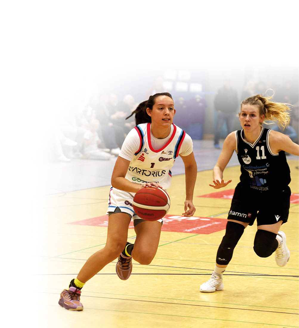 Eine Spielszene aus einem Basketballspiel in der Halle: Eine Frau im wei en Trikot dribbelt den Ball nach vorne, w hrend eine verteidigende Frau im schwarzen Trikot dicht folgt. Im Hintergrund sind Zuschauende auf der Trib ne sowie weitere Hallenelemente zu sehen.