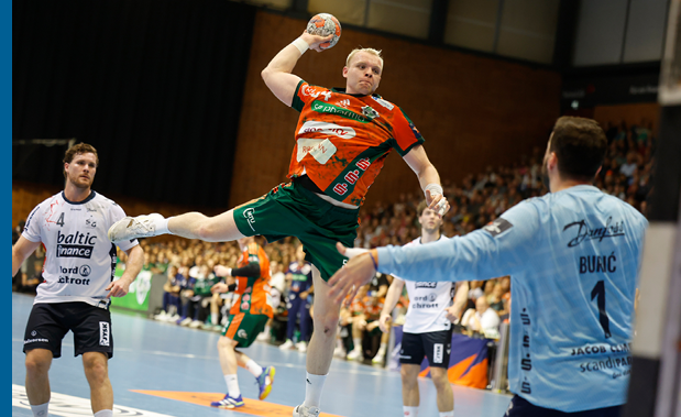 Ein Handballspieler in orange-gr nem Trikot springt w hrend eines Spiels hoch in die Luft und wirft auf das Tor. Vor ihm versucht der Torwart im hellblauen Trikot den Ball abzuwehren, w hrend weitere Spieler und Zuschauer im Hintergrund zu sehen sind.