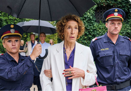 “Mama ist die Best(i)e": Zwei Polizisten f hren Gloria Almeda (Adele Neuhauser), die sich in ihrer Mitte befindet. Eine der beiden Polizisten tr gt einen schwarzen Regenschirm, den sie ber Gloria h lt. Glorias Blick dr ckt Verzweiflung aus. Im Hintergrund stehen die Kommissarin und die Familie Glorias.