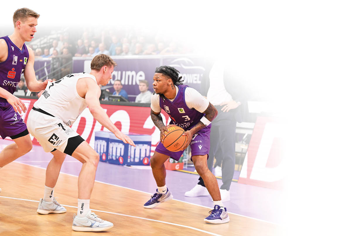 Basketballspiel in einer vollen Halle: Ein Spieler im lila Trikot dribbelt den Ball und wird eng von einem Spieler im wei en Trikot verteidigt, w hrend ein weiterer Spieler in Lila zur Unterst tzung bereitsteht und eine Person in Zivilkleidung am Spielfeldrand zuschaut.