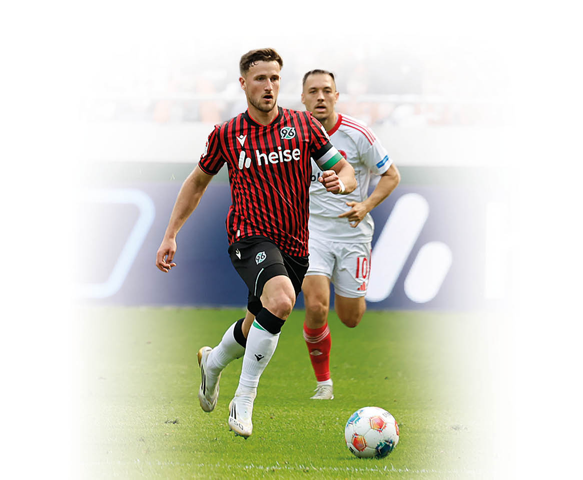 Fu ballszene auf dem Spielfeld: Ein Spieler im rot-schwarz gestreiften Trikot mit „heise“-Aufdruck sprintet mit dem Ball nach vorne, dicht verfolgt von einem Gegenspieler in wei em Trikot; im Hintergrund ist eine gut besetzte Trib ne zu sehen.