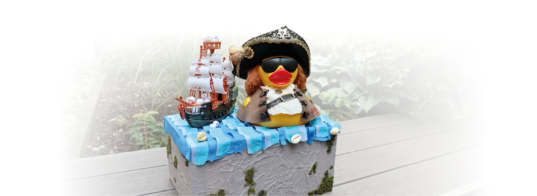 Eine kleine dekorative Szene zeigt eine Ente mit Piratenhut und Sonnenbrille neben einem Modellschiff auf einer mit blauen Wellen verzierten Box im Freien. Beschreibung von KI generiert