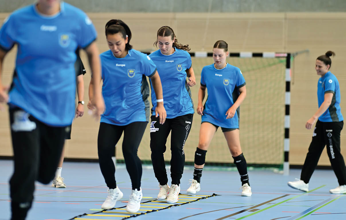 Mehrere Erwachsene in blauen Sportshirts und schwarzen Hosen trainieren in einer Halle mit einem Koordinationsleiter auf dem Boden vor einem Handballtor. Beschreibung von KI generiert