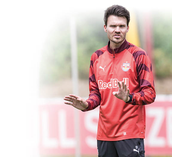 Eine Person mit kurzem Haar tr gt ein rotes Sporttrikot mit Red Bull-Logo und gestikuliert auf einem Fu ballfeld vor einem unscharfen Hintergrund mit der Aufschrift Salzburg. Beschreibung von KI generiert