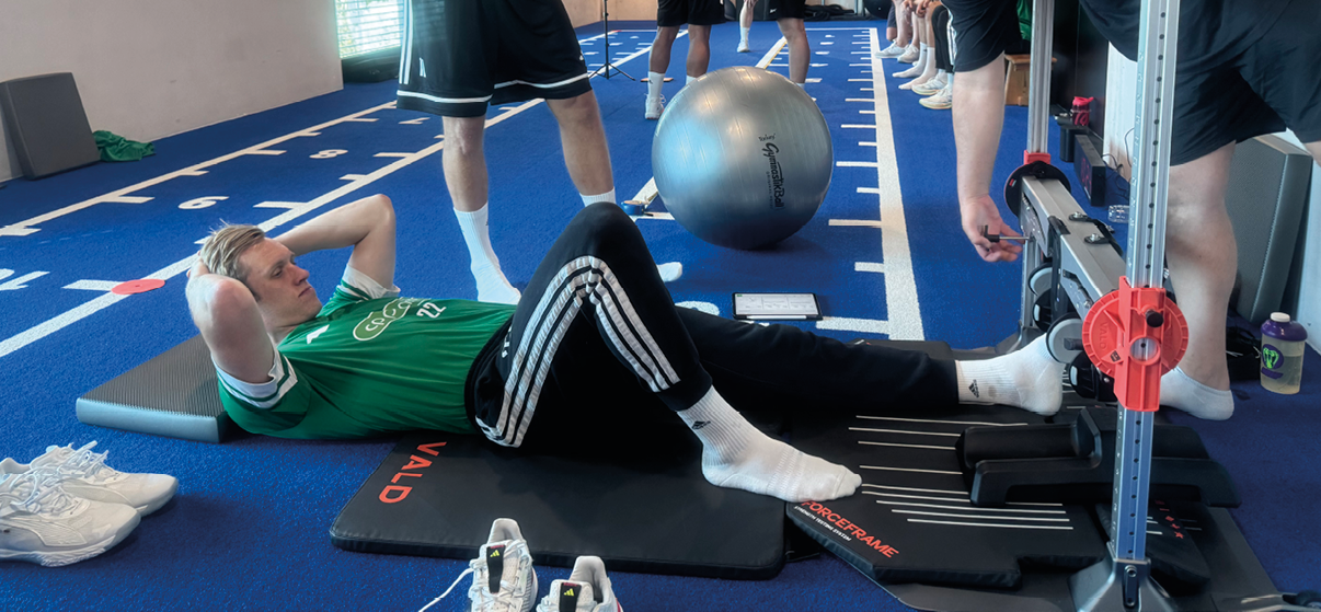 Mehrere Personen in Sportkleidung trainieren in einem Fitnessraum mit blauen Bodenmatten, wobei eine Person auf einer Matte liegt und eine andere Person an einem Trainingsger t arbeitet. Beschreibung von KI generiert