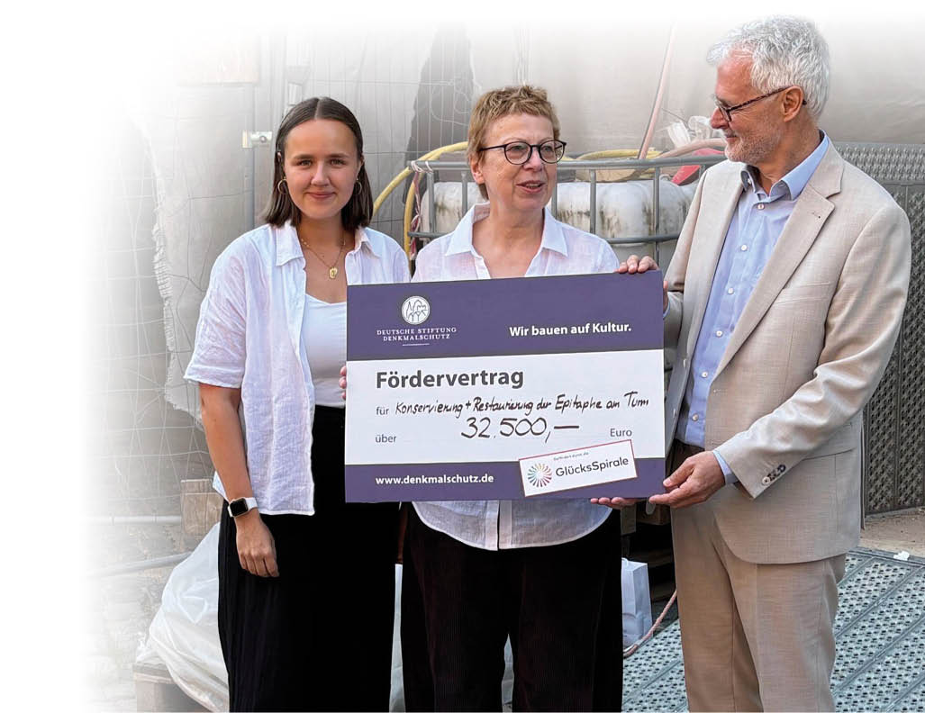 Drei Personen stehen zusammen und halten einen gro en symbolischen Scheck ber eine F rderung f r den Erhalt eines historischen Geb udes.  Beschreibung von KI generiert
