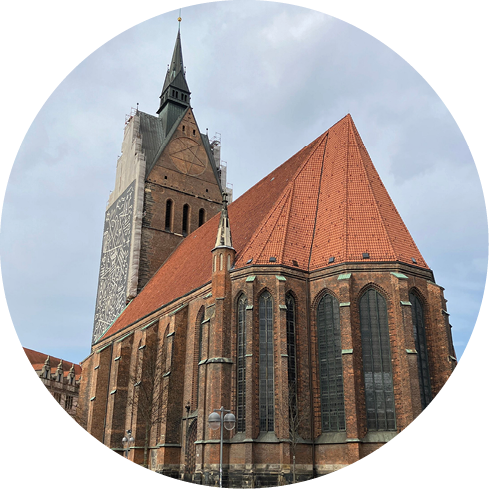 Eine gotische Backsteinkirche mit hohen spitzbogigen Fenstern und einem steilen roten Ziegeldach unter einem bew lkten Himmel. Beschreibung von KI generiert