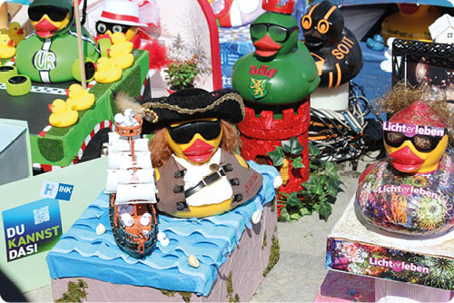 Mehrere bunte Gummienten sind in einer kreativen Szene mit verschiedenen Kost men und Dekorationen aufgestellt, darunter eine Ente als Pirat mit einem kleinen Schiff auf blauem Wasser und eine andere mit Sonnenbrille und einem bunten Pullover. Beschreibung von KI generiert
