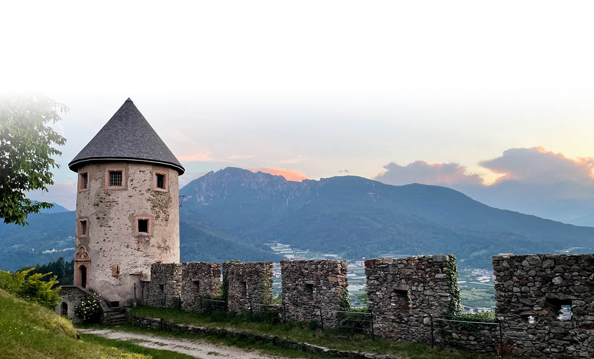 Eine alte Steinburg mit einem runden Turm steht auf einem H gel mit Blick auf bewaldete Berge und einen bew lkten Himmel bei Sonnenuntergang. Beschreibung von KI generiert