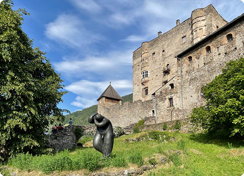 Eine mittelalterliche Burg steht auf einem H gel mit gr ner Wiese und B umen, davor befindet sich eine moderne abstrakte Skulptur und einige Personen sitzen auf einer Steinbr cke. Beschreibung von KI generiert