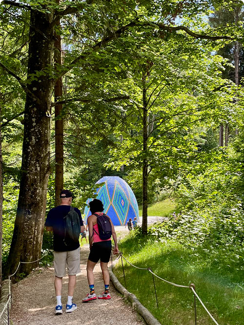 Zwei Personen mit Rucks cken gehen auf einem Waldweg entlang, w hrend im Hintergrund eine bunte, kuppelf rmige Struktur zwischen den B umen steht. Beschreibung von KI generiert