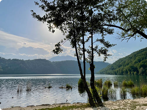 Ein ruhiger See mit B umen am Ufer und H geln im Hintergrund unter einem bew lkten Himmel mit Sonnenlicht. Beschreibung von KI generiert